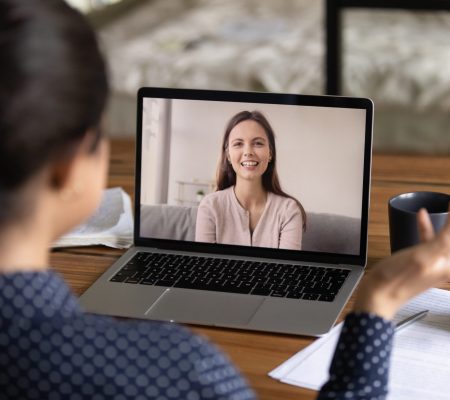 Happy young millennial colleagues having virtual talk, discussing work project. Teacher training student via video call. Women teaching and studying language online. Screen display view over shoulder